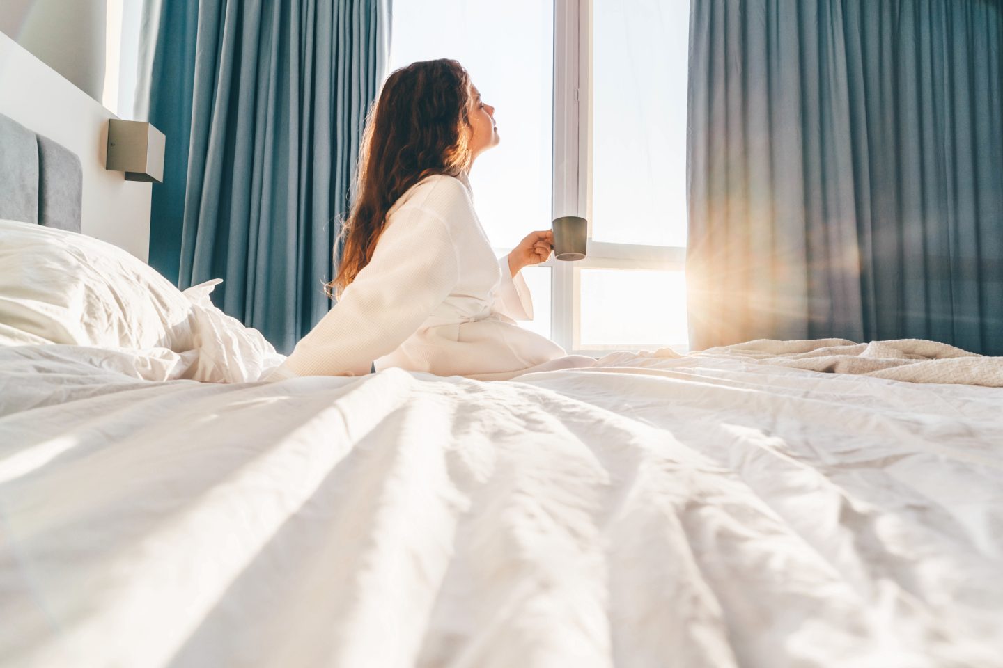 Woman in a white robe on her bed with a mug in morning light, dealing with dry, itchy menopause skin